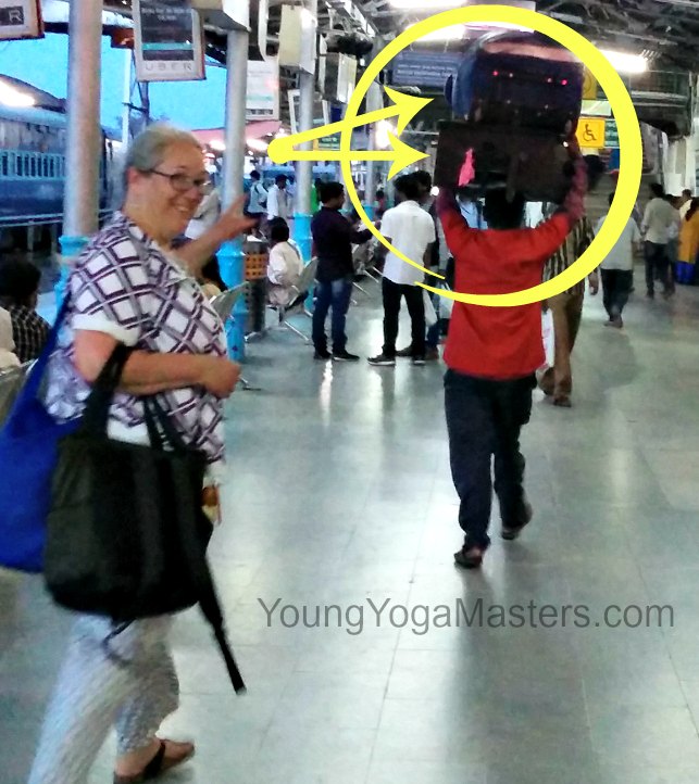 a yoga teacher in india with two suitcases being carried on the head of a porter through the train station