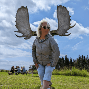 Aruna stands outdoors with a blue sky and green grass field. She is standing in front of moose antlers behind her head and making a silly pose that makes her look like she has antlers. 